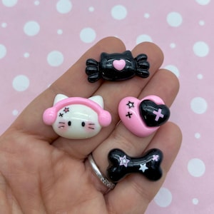 May include: A set of four black and pink resin charms featuring a cat with headphones, a candy, a heart, and a bone. Each charm has a white star and a cross design.
