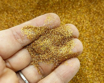 May include: A close-up of a hand holding a pile of fine gold glitter. The glitter is very sparkly and has a metallic sheen.