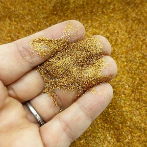 Op de afbeelding: Een close-up van een hand die een hoop fijne gouden glitter vasthoudt. De glitter is erg glinsterend en heeft een metallic glans.