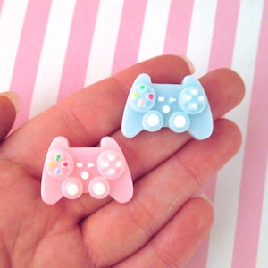 May include: Two miniature plastic game controllers, one light blue and one pink, with colorful buttons. The controllers are held in a hand against a pink and white striped background.