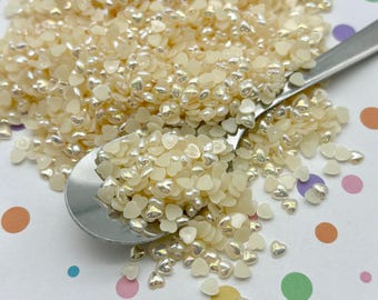 May include: A close-up of a spoon filled with iridescent white heart-shaped sprinkles. The sprinkles are scattered on a white surface with colorful polka dots.