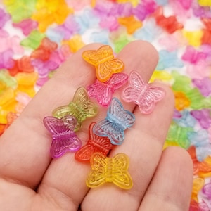 Puede incluir: Una colección de cuentas translúcidas en forma de mariposa, en varios tonos como naranja, rosa, verde, azul, rojo y amarillo. Las cuentas están dispersas sobre una superficie con más cuentas de mariposa en el fondo.