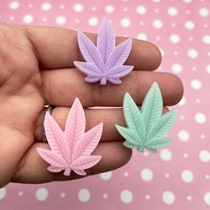 May include: Three pastel-colored resin cannabis leaf charms. The leaves are in shades of light purple, pink, and green.