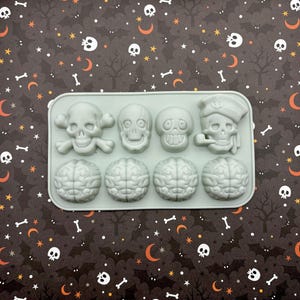 Könnte beinhalten: Eine hellgraue Silikonform mit Schädel- und Gehirnformen. Die Form enthält einen Totenkopf mit gekreuzten Knochen, drei verschiedene Schädeldesigns und vier Gehirnformen. Die Form befindet sich auf einem gemusterten Hintergrund mit Totenköpfen, Knochen und Monden.