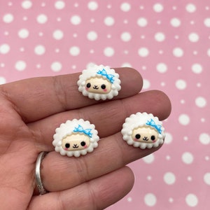 May include: Three white resin sheep charms with blue bows. The sheep have a cute cartoon face with black eyes and a pink nose.