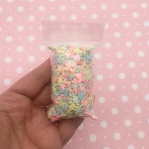 May include: A small clear plastic bag filled with pastel colored heart-shaped sprinkles. The sprinkles are in shades of pink, blue, yellow, and green.