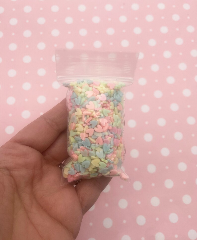 May include: A small clear plastic bag filled with pastel colored heart-shaped sprinkles. The sprinkles are in shades of pink, blue, yellow, and green.