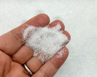 May include: A pile of small, translucent, crystalline white granules resting in an open palm. The particles are irregular in shape and size, reflecting light with a sparkling appearance, resembling coarse salt, sugar, or decorative craft sand.