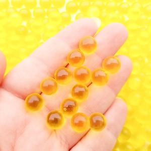 May include: A hand holding several small, translucent, amber-coloured spheres. The spheres are scattered across the palm and fingers, with a background of similar spheres. The spheres appear to be gel-like.