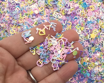 Puede incluir: Un puñado de confeti en forma de letras iridiscente pastel. El confeti está hecho de plástico y tiene un acabado holográfico. El confeti es de varios colores, incluyendo rosa, azul, amarillo y morado.