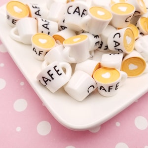 May include: A white plate filled with miniature ceramic coffee cups. The cups are decorated with the word "Cafe" and a small heart. The cups are on a pink background with white polka dots.