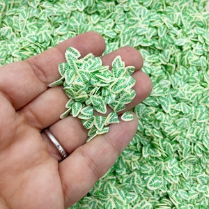May include: A collection of small, leaf-shaped polymer clay pieces. The leaves are mainly light green with white and darker green details, giving a natural look. The background is filled with similar clay pieces.