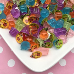 May include: A collection of colorful, individually packaged jelly cups in shades of orange, blue, purple, green, and pink. The jelly cups are arranged on a white square plate, with a pink polka dot background.