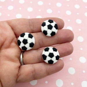 May include: Three white and black soccer ball shaped polymer clay charms. The charms are approximately 1 inch in diameter.