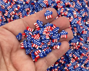 Op de afbeelding: Een hand die een stapel kleine, rood-wit-blauwe, stervormige polymer clay charms vasthoudt. De charms hebben de vorm van een ster met een rood-wit gestreept patroon.