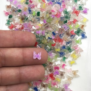May include: A large assortment of colorful glitter butterfly charms. The charms are made of clear resin with glitter embedded in the resin. The butterflies are in various colors, including pink, purple, blue, green, and yellow. The charms are small, about the size of a fingernail.