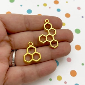 Op de afbeelding: Twee gouden metalen honingraatbedels. De bedels zijn in de vorm van een honingraat met zes cellen. De bedels liggen op een witte achtergrond met kleurrijke stippen.