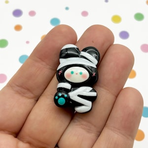May include: A small black and white mummy-wrapped bear with blue eyes and a turquoise paw print. The bear is made of polymer clay and is a cute Halloween decoration.