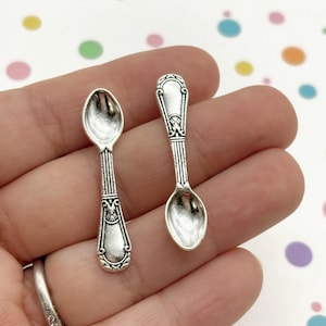 May include: Two small, silver spoons with decorative handles and oval bowls. The spoons are held in a hand against a background of colourful polka dots. The spoons have a vintage or antique appearance.