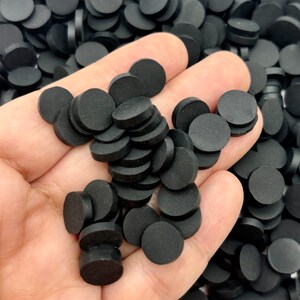 May include: A handful of small, black, circular objects. The objects are stacked and appear to be made of a matte material. The background is filled with similar objects, suggesting a large quantity of the same item.