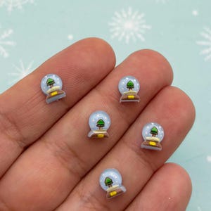 Puede incluir: Cinco encantos de bola de nieve en miniatura con una cúpula de vidrio azul, nieve blanca y un árbol de Navidad verde en el interior. La base de cada encanto es marrón.