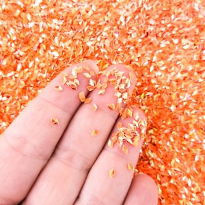 May include: A close-up of a hand holding orange iridescent glitter. The glitter is in the shape of small diamonds.
