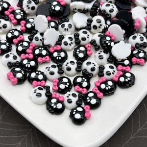 Può includere: Una collezione di abbellimenti a forma di teschio bianchi e neri con fiocchi rosa. I pezzi decorativi sono sparsi su una superficie bianca, con uno sfondo scuro. I teschi hanno occhi bianchi e sono adatti per progetti di artigianato.