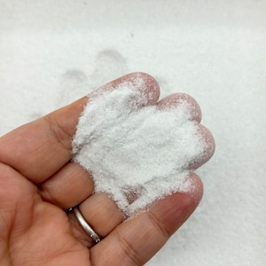 Puede incluir: Primer plano de una mano sosteniendo un montón de purpurina blanca. La purpurina es muy fina y tiene un ligero brillo.