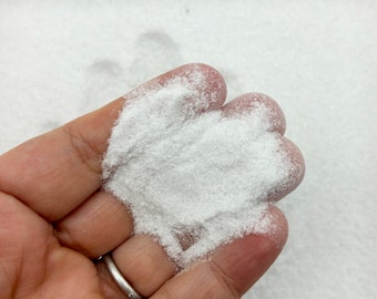 May include: A close-up of a hand holding a pile of white glitter. The glitter is very fine and has a slight sparkle to it.