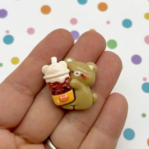 Op de afbeelding: Een bruine berenfiguur die een bruin en wit boba-theekopje vasthoudt met een wit deksel en een gele rietje.
