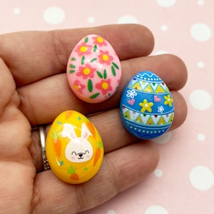 May include: Three hand-painted Easter eggs. One egg is pink with pink flowers, one is blue with yellow and white geometric designs, and one is yellow with a white bunny and orange carrots.