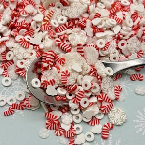 May include: A spoon filled with white and red candy sprinkles, including candy canes, snowflakes, and donut shapes. The sprinkles are scattered on a light blue background with white snowflake designs.