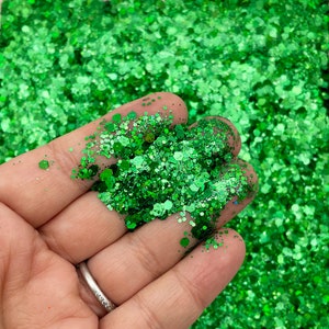 May include: A close-up of green glitter with a variety of sizes and shapes. The glitter is being held in a hand.