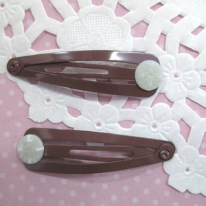 May include: Two brown plastic hair clips with white pearl-like accents. The clips are on a pink background with white polka dots.