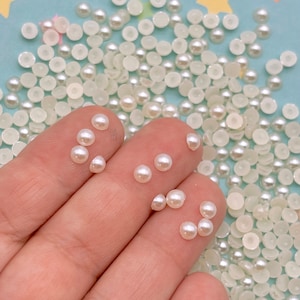 May include: A close-up of a hand holding a handful of small, white pearl-like beads. The beads are scattered on a light blue surface with yellow stars.