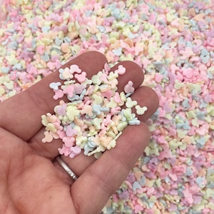 May include: A close-up of pastel-colored Mickey Mouse head sprinkles. The sprinkles are in a variety of colors, including pink, blue, yellow, and white.