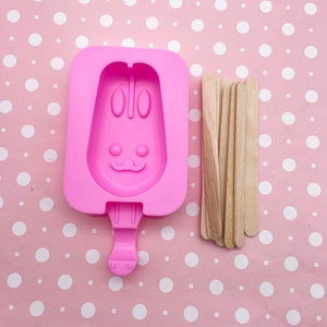 Op de afbeelding: Roze siliconen mal in de vorm van een konijnenlolly met een stokje. De mal is op een roze en wit gestippelde achtergrond met houten lollystokjes.