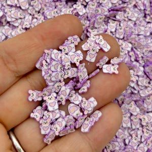May include: A close-up of a hand holding a pile of small, white and purple resin bear charms. The bears have a heart on their chest and are perfect for adding to slime, resin, or other crafts.