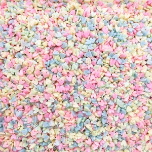 May include: A close-up image of pastel-colored heart-shaped sprinkles. The sprinkles are a mix of light blue, pink, yellow, and white.