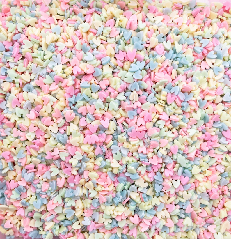 May include: A close-up image of pastel-colored heart-shaped sprinkles. The sprinkles are a mix of light blue, pink, yellow, and white.