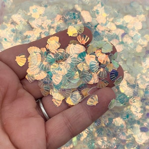 May include: Iridescent seashell-shaped glitter in various sizes. The glitter has a pearlescent sheen, reflecting colors of gold, blue, and green. The glitter is held in a hand, with many more pieces in the background.