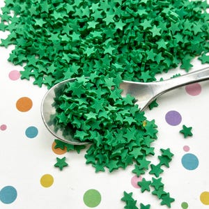 May include: A close-up of a spoon filled with green star-shaped sprinkles. The sprinkles are scattered on a white surface with colorful polka dots.