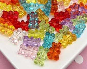 May include: A collection of colorful gummy bear shaped beads in various shades of red, orange, yellow, green, blue, purple, and clear. The beads are scattered on a white plate.