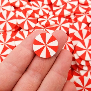 May include: A close-up of a red and white candy cane-shaped embellishment. The embellishment is made of polymer clay and is about 1 inch in diameter. The embellishment is being held by a hand against a background of many similar embellishments.