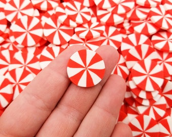 May include: A close-up of a red and white candy cane-shaped embellishment. The embellishment is made of polymer clay and is about 1 inch in diameter. The embellishment is being held by a hand against a background of many similar embellishments.