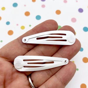 Può includere: Due fermagli per capelli in metallo bianco. Ogni fermaglio ha tre fessure orizzontali e un'estremità arrotondata. I fermagli sono tenuti in mano su uno sfondo di carta bianca con pois colorati.