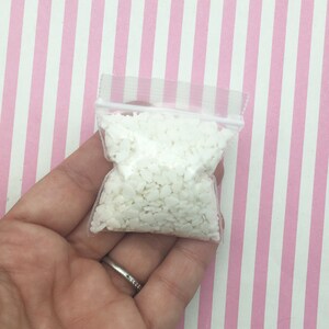 May include: A small, clear plastic bag filled with numerous white, heart-shaped wax beads. The bag is held in a hand against a backdrop of pink and white striped paper. The beads appear to be for crafting or candle making.