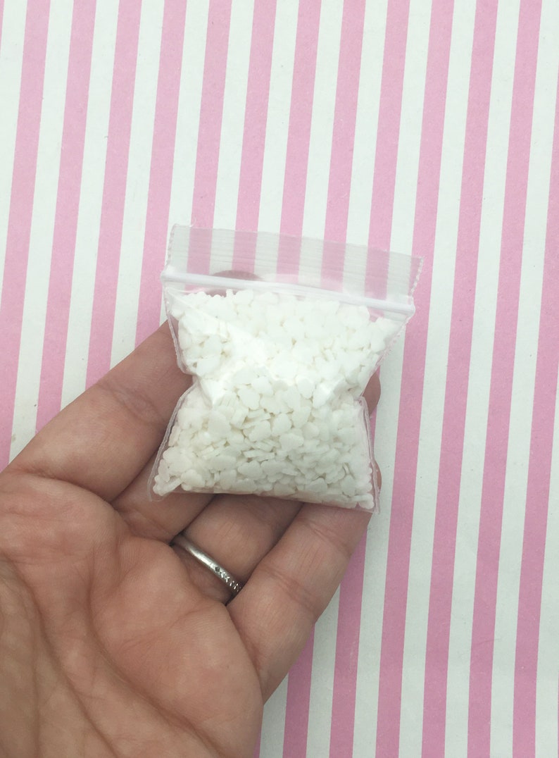 May include: A small, clear plastic bag filled with numerous white, heart-shaped wax beads. The bag is held in a hand against a backdrop of pink and white striped paper. The beads appear to be for crafting or candle making.