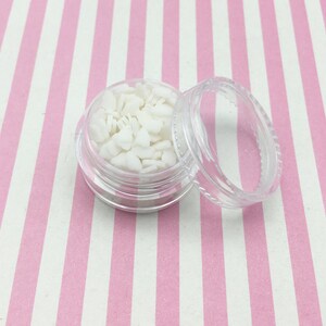 May include: A small, clear plastic jar filled with white bunny-shaped confetti. The jar's lid is off, revealing the contents. The background is a pink and white striped pattern.