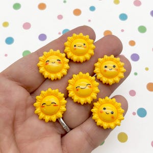Könnte beinhalten: Eine Sammlung von sieben gelben, sonnenblumenförmigen Verzierungen mit lächelnden Gesichtern. Jede Sonnenblume hat ein detailliertes Blütenblattdesign und einen fröhlichen Ausdruck. Die Verzierungen werden in einer Hand vor einem gepunkteten Hintergrund gehalten.